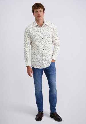 Camicia bianca a bottoni con pois neri, maniche lunghe e vestibilità casual, abbinata a jeans blu e scarpe scure.