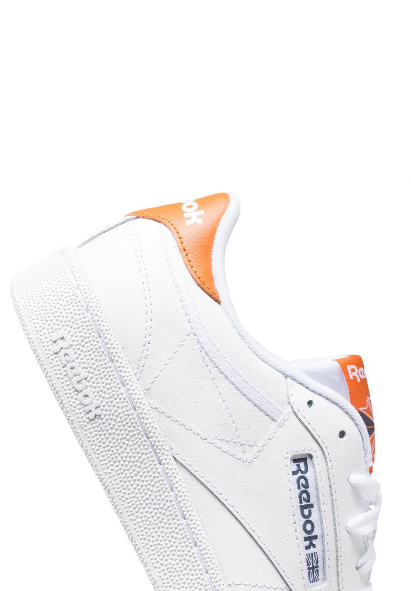 Zapatilla blanca de cuero con acentos naranjas en el talón, suela de goma texturizada y logotipo de Reebok en el lateral y la lengua. Diseño minimalista.