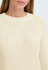Beige strikket genser med ribbestrikket rund hals og et teksturert mønster. Har raglanermer og en avslappet passform, egnet for lag-på-lag.