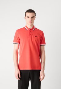 Rød polo-skjorte laget av bomull, med klassisk krage, tre knapper og kontrasterende stripete detaljer på ermene. Svart logo på brystet.