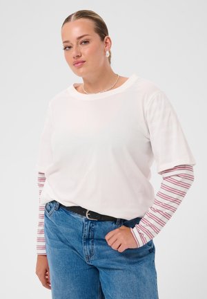 Femme aux cheveux plaqués en arrière portant une chemise blanche à manches courtes par-dessus un top à manches longues rayé rose et blanc et un jean bleu avec une ceinture noire.