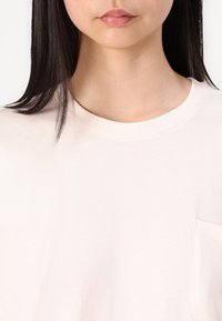 Svetle růžové bavlněné tričko s kulatým výstřihem a malou kapsou na levé straně. Měkká textura a minimalistický design.