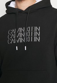 Felpa nera con logo bianco di Calvin Klein stampato sul davanti. Caratterizzata da cappuccio con cordoncino e tasca frontale. Texture del tessuto liscia.