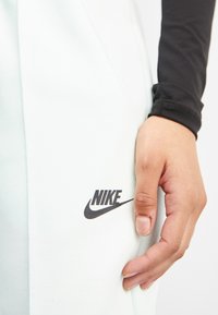 Licht mintgroene Nike joggers met een zwart logo op het onderbeen en een hand die op de stof rust. Zachte textuur en slank ontwerp.