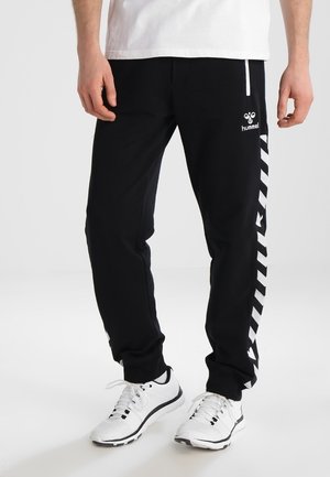 Mężczyzna ubrany w czarne spodnie dresowe Hummel z białymi paskami w kształcie litery V po bokach oraz białe buty sportowe, stojący na jednolitym tle.