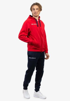 Felpa rossa con zip e cordini blu, abbinata a pantaloni da jogging blu navy con accenti di logo bianchi. Sneakers bianche completano l'outfit.