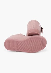 Zapatillas rosa tipo slip-on con parte superior de tela texturizada, suela plana de goma y detalle de botón en el lateral. La suela tiene un patrón de agarre estriado.