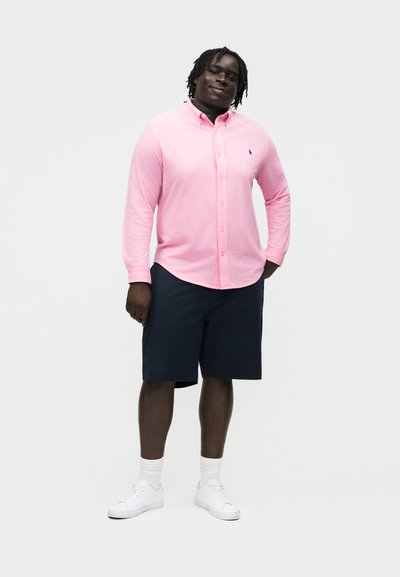 Mann som smiler med hendene i lommene, iført en rosa skjorte med knapper, marineblå shorts, hvite sokker og hvite joggesko, stående mot en ensfarget bakgrunn.