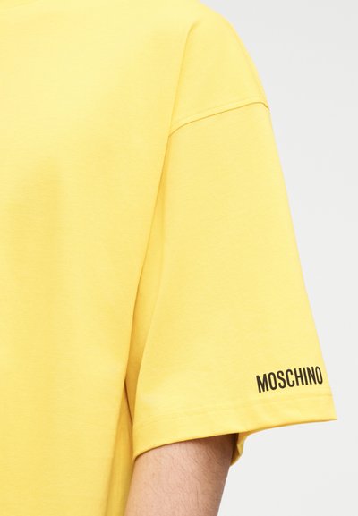 Κίτρινη βαμβακερή μπλούζα με χαλαρή εφαρμογή, κοντά μανίκια και μαύρο λογότυπο «Moschino» τυπωμένο στο αριστερό μανίκι. Ομαλή υφή.