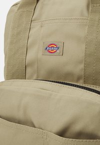 Kaki spalva kuprinė, pagaminta iš tvirto audinio, su juodu užtrauktuku, priekinėmis kišenėmis ir ryškiu Dickies logotipu šone.