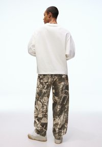 Bílá košile s dlouhým rukávem, volný střih, doplněná kalhotami s maskáčovým vzorem a pohodlným střihem. Tenisky s béžovými detaily. Neutrální pozadí.