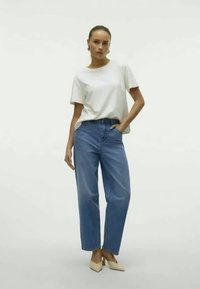 T-shirt branca de manga curta, jeans azul de cintura alta e sapatos de salto apontados bege. Modelo está de pé com uma mão na cintura, contra um fundo neutro.