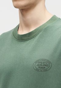 Άτομο που φοράει πράσινο t-shirt με στρογγυλή λαιμόκοψη και μικρό μαύρο οβάλ λογότυπο με την επιγραφή "Jack & Jones Premium" στο αριστερό μέρος του στήθους.