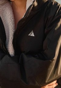 Zwarte jas met ritssluiting en een heerlijk zachte, witte binnenkant, voorzien van een zilveren driehoekig logo op de linkerborst en brede, zachte mouwen.