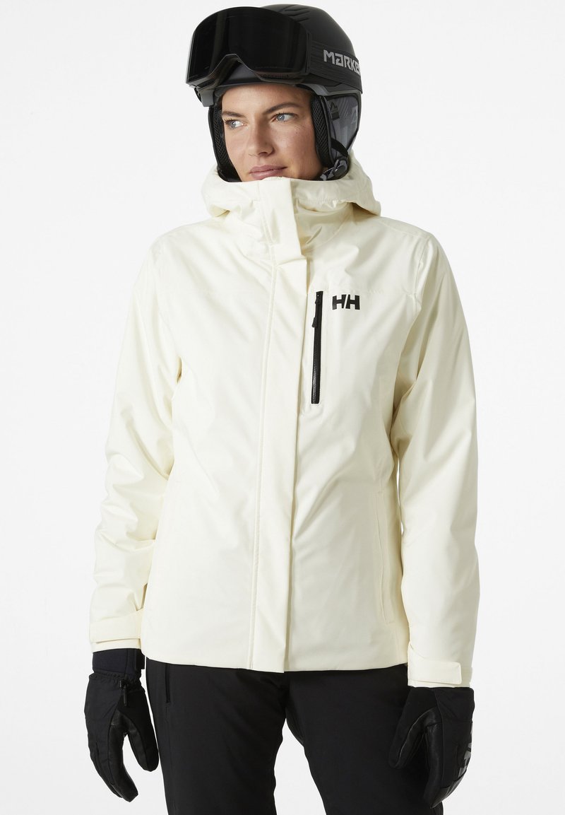 Witte waterdichte skijas met een hoge kraag, zwarte ritszak en logo. Draagt zwarte handschoenen en een zwarte skihard.}}