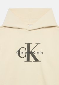 Béžová mikina s kapucí, která má velký černý design loga se slovy "CK" a "CalvinKlein". Měkká látka s prostornou, skládací kapucí.