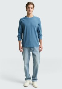 Blaut long-sleevi T-Shirt mit rundem Halsausschnitt und gerippten Bündchen, kombiniert mit hellblauen Jeans und weißen Sneakers. Einfaches Design, lässige Passform.