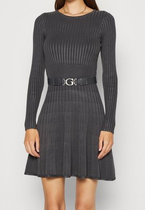 Frau trägt ein dunkelgraues, geripptes Strickkleid mit langen Ärmeln, ausgestelltem Rock und einem schwarzen Gürtel mit silberner G-Schnalle an der Taille.