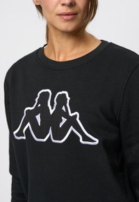 Zwarte sweatshirt met een witte geborduurde logo van twee zittende figuren. Ronde hals en zachte, gestructureerde stof.