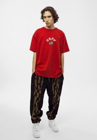 Rode katoenen t-shirt met witte geborduurde tekst en tankafbeelding. Zwarte broek met een gouden abstract patroon. Witte sneakers maken de look compleet.