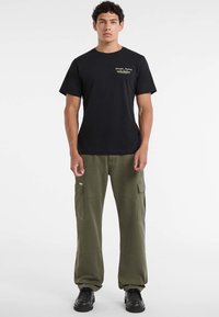 Młody mężczyzna stoi w czarnym t-shircie Guess Jeans, oliwkowych spodniach cargo i czarnych butach na jednolitym białym tle.
