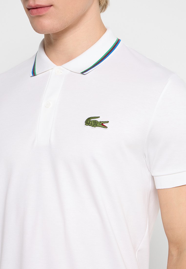T-shirt polo blanche avec patte de boutonnage, col côtelé arborant des rayures vertes, bleues et rouges, et un logo crocodile brodé en vert.