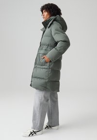 Giacca trapuntata in verde tenue, con cappuccio, chiusura a zip e tasche laterali. Abbinata a jeans a gamba larga grigi e sneakers bianche con dettagli neri.