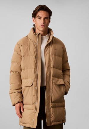 Ung mand iført en beige foret vinterjakke med lommer foran og høj krave, stående mod en ensfarvet grå baggrund.
