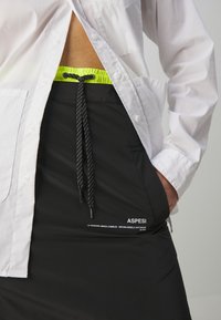 Jupe noire avec une taille à cordon, dotée d'une doublure intérieure vert néon et d'un cordon rayé noir et blanc. Comprend un détail de logo de marque blanc.