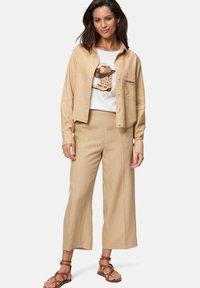 Beige knoophemd over een wit T-shirt met opdruk, gecombineerd met beige wijde broek. Bruine sandaaltjes met banden maken de outfit af.