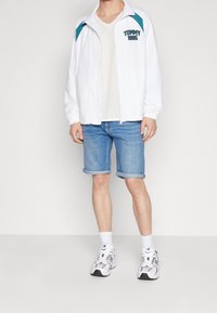 Chaqueta ligera blanca con detalles en color azul verdoso y "TOMMY 1985" impreso en el pecho, combinada con pantalones cortos de mezclilla azul con dobladillo enrollado y zapatillas deportivas.