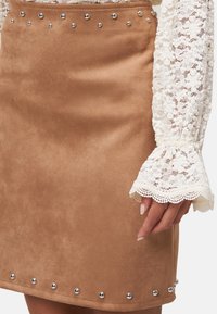 Jupe en faux daim marron avec des clous en argent le long de l'ourlet. Présentée avec une manche en dentelle blanche texturée qui souligne le détail.