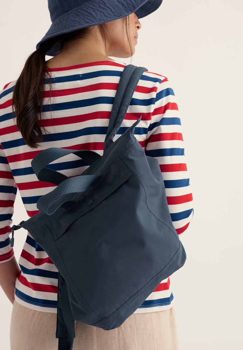 Personne portant une chemise rayée rouge, blanche et bleue et un chapeau bob bleu marine, portant un sac à bandoulière bleu marine sur un fond uni.