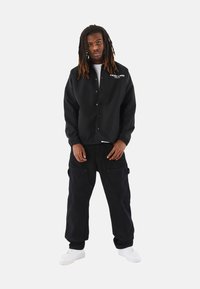 Chaqueta negra con botones a presión y puños elásticos, combinada con pantalones cargo negros que tienen bolsillos, y zapatillas blancas.