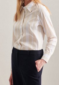 Woman with beige and white vertical striped button-up shirt tucked into black pants, standing with one hand in pocket.
