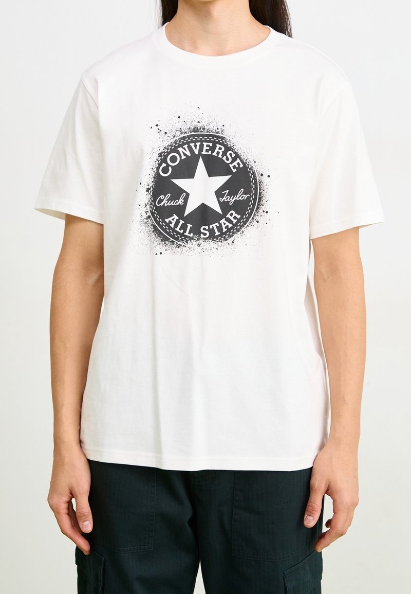 Personne portant un t-shirt blanc avec un logo Converse All Star Chuck Taylor noir et un pantalon foncé, visible du cou jusqu'aux hanches.