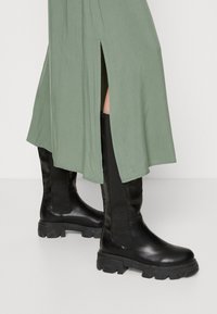 Bottes noires en cuir, montantes jusqu'au genou, avec des côtés texturés et des semelles en caoutchouc épaisses. Portées avec une jupe verte présentant une fente sur le côté.