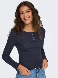 Tmavomodrý rebrovaný top s dlhým rukávom, okrúhlym výstrihom, zapínaním na tri gombíky a vlnitým lemom, v kombinácii s džínsami s vysokým pásom.