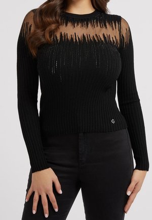 Femme portant un pull noir ajusté à manches longues avec une partie supérieure de la poitrine transparente et des rayures verticales décoratives, associé à un pantalon noir.