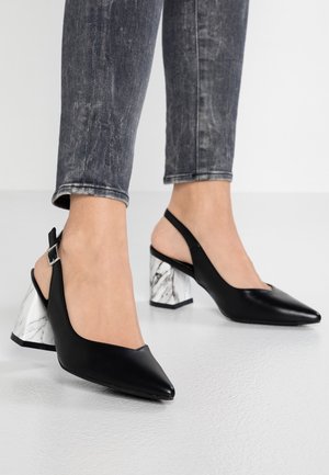 Stopy w czarnych sandałkach na obcasie typu slingback z białymi obcasami w marmurowy wzór oraz w szarych, przetartych obcisłych dżinsach na białej podłodze.