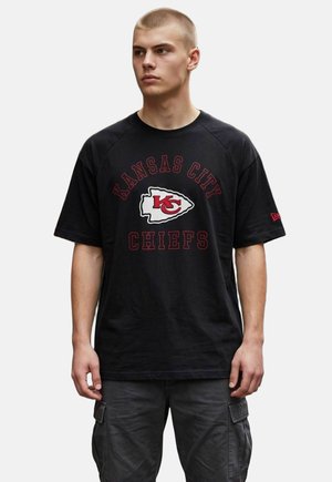 Młody mężczyzna z krótkimi blond włosami, ubrany w czarną koszulkę Kansas City Chiefs i ciemne spodnie typu cargo, patrzy w bok.