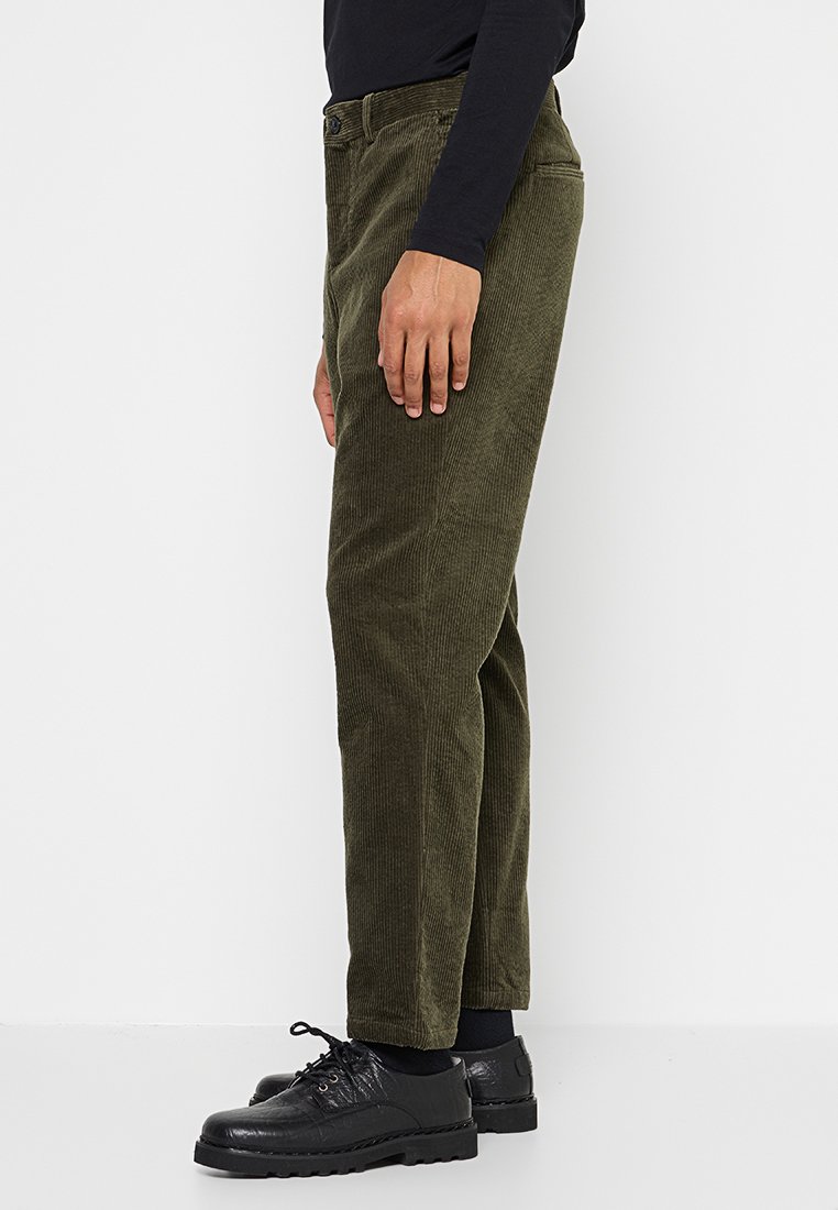 Olivgrüne Cordhosen mit geradem Bein, Knopfverschluss und seitlichen Taschen, kombiniert mit schwarzen Schuhen. Strukturierter Stoff sichtbar.