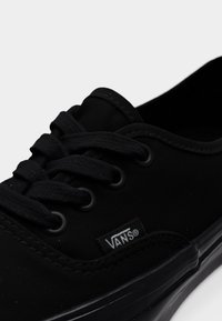 Μαύρα πάνινα παπούτσια με κορδόνια, με λαστιχένια σόλα, εμφανή ραφή και μικρή λευκή ετικέτα με λογότυπο στο πλάι.