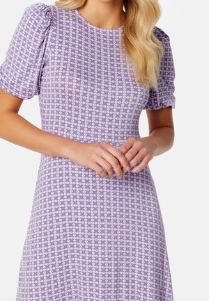 Kobieta w lawendowej sukience z białym drobnym wzorem kwiatowym, z krótkimi bufkami na rękawach, delikatnie trzymająca dłonie splecione na talii.