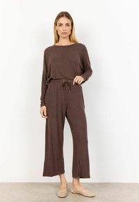 Bruine langemouwtop en bijpassende wijd-uitlopende broek met een trekkoord in de taille. De stof heeft een zachte textuur. Beige, versierde schoenen maken de outfit compleet.