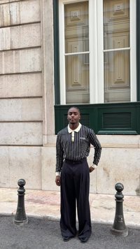 Gestreiftes, schwarz-weißes Langarmshirt mit einem cremefarbenen Kragen, kombiniert mit lockeren schwarzen Hosen, stehend vor einer strukturierten Steinwand.