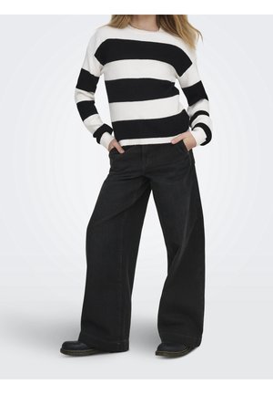 Personne portant un pull rayé noir et blanc avec un pantalon noir à jambes larges et des chaussures noires, debout les mains dans les poches.