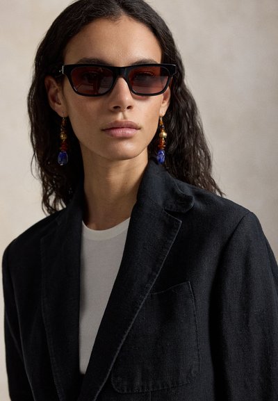 Mlada žena s dugom kovrčavom kosom, nosi tamne pravokutne sunčane naočale, šarene višeće naušnice, crni sako i bijelu majicu, na neutralnoj pozadini.