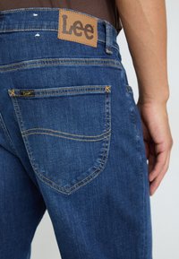 Jeanșii din denim de culoare albastru închis au o etichetă din piele inscripționată cu "Lee" pe talie și o cusătură galbenă contrastantă pe buzunarele din spate.
