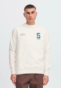 Hanorac crem cu manșete și tiv cu nervuri, având brodat pe piept un „S” verde și text. Modelul poartă pantaloni maro. Design simplu.